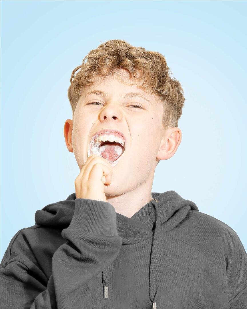 Ein Junge mit lockigem Haar und dunklem Kapuzenpulli setzt sich vor einem hellblauen Hintergrund mit der Hand eine durchsichtige Zahnspange in den Mund.