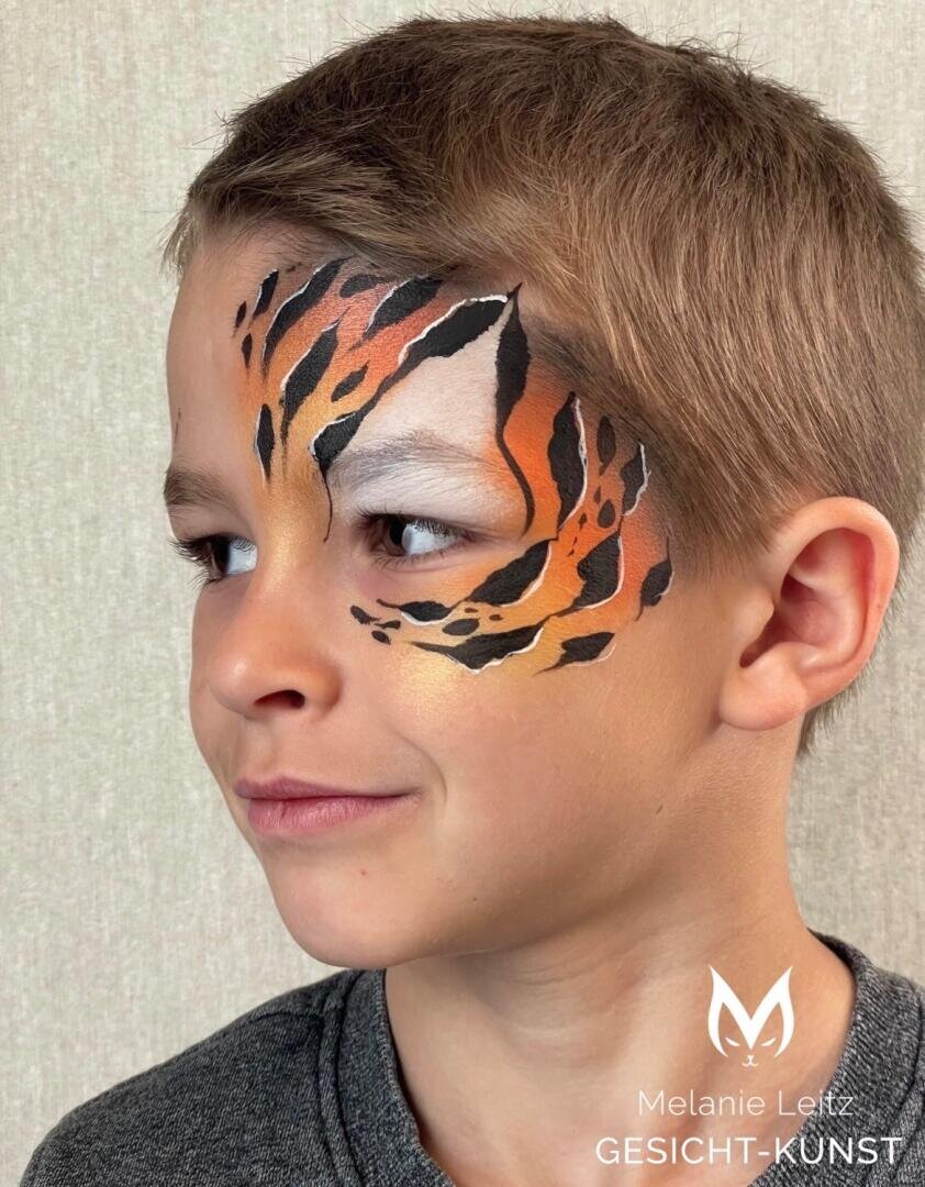 Das Gesicht eines Jungen mit kurzen braunen Haaren ist teilweise mit einem orangefarbenen, schwarzen und weißen Tigermotiv bemalt, das sich auf die Augen und die Stirn konzentriert. Der Hintergrund ist hell und schlicht. Das Wasserzeichen lautet Melanie Leitz GESICHT-KUNST.