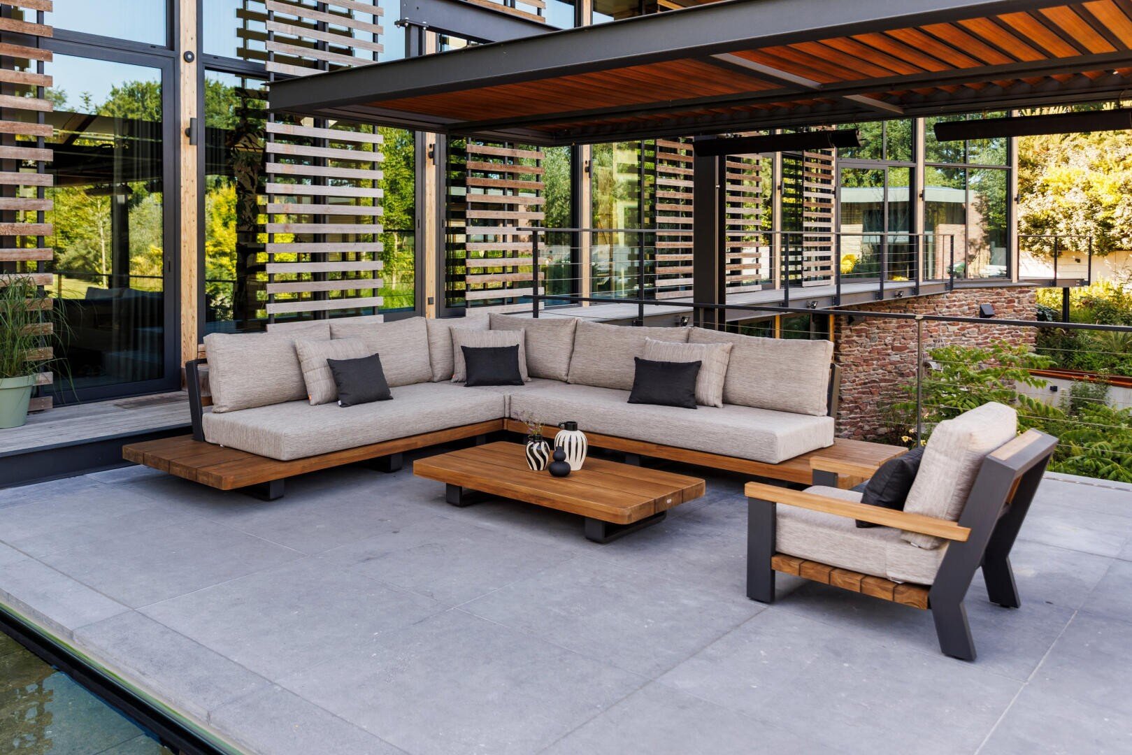 Moderne Außenterrasse mit einem großen L-förmigen beigen Sofa, einem passenden Sessel, einem hölzernen Couchtisch, dekorativen Vasen und einer Pergola über dem Dach. Im Hintergrund sind Glaswände und Grünflächen zu sehen.