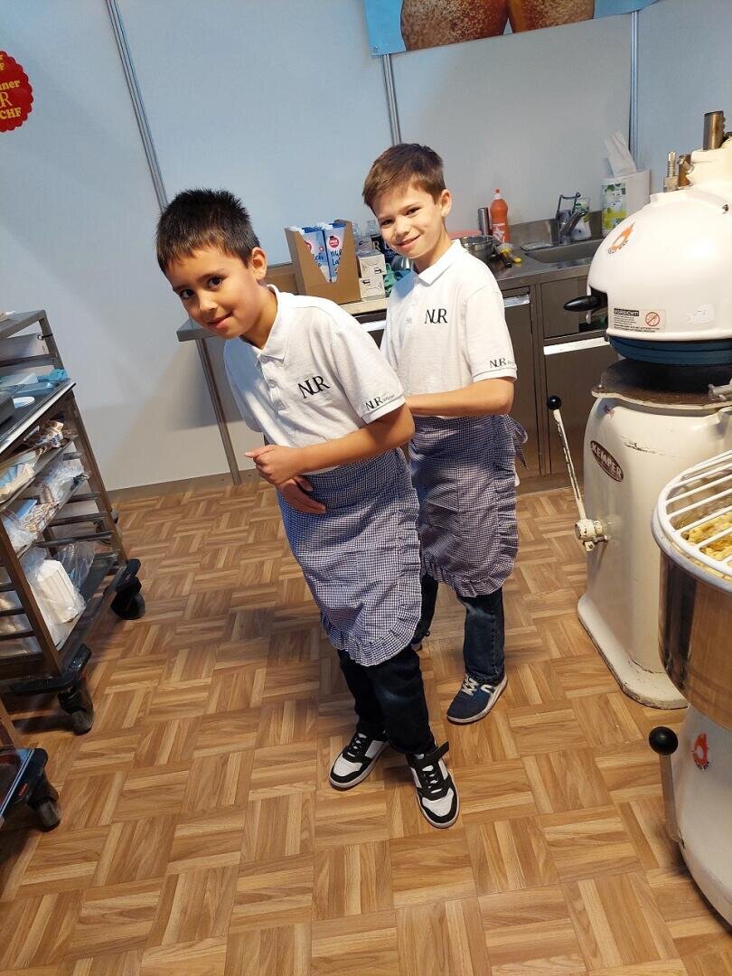 Zwei Jungen in passenden weißen Poloshirts und blauen Schürzen stehen Rücken an Rücken in einer Küche und lächeln spielerisch. Um sie herum sind auf einem Holzboden Backgeräte und -zubehör zu sehen.