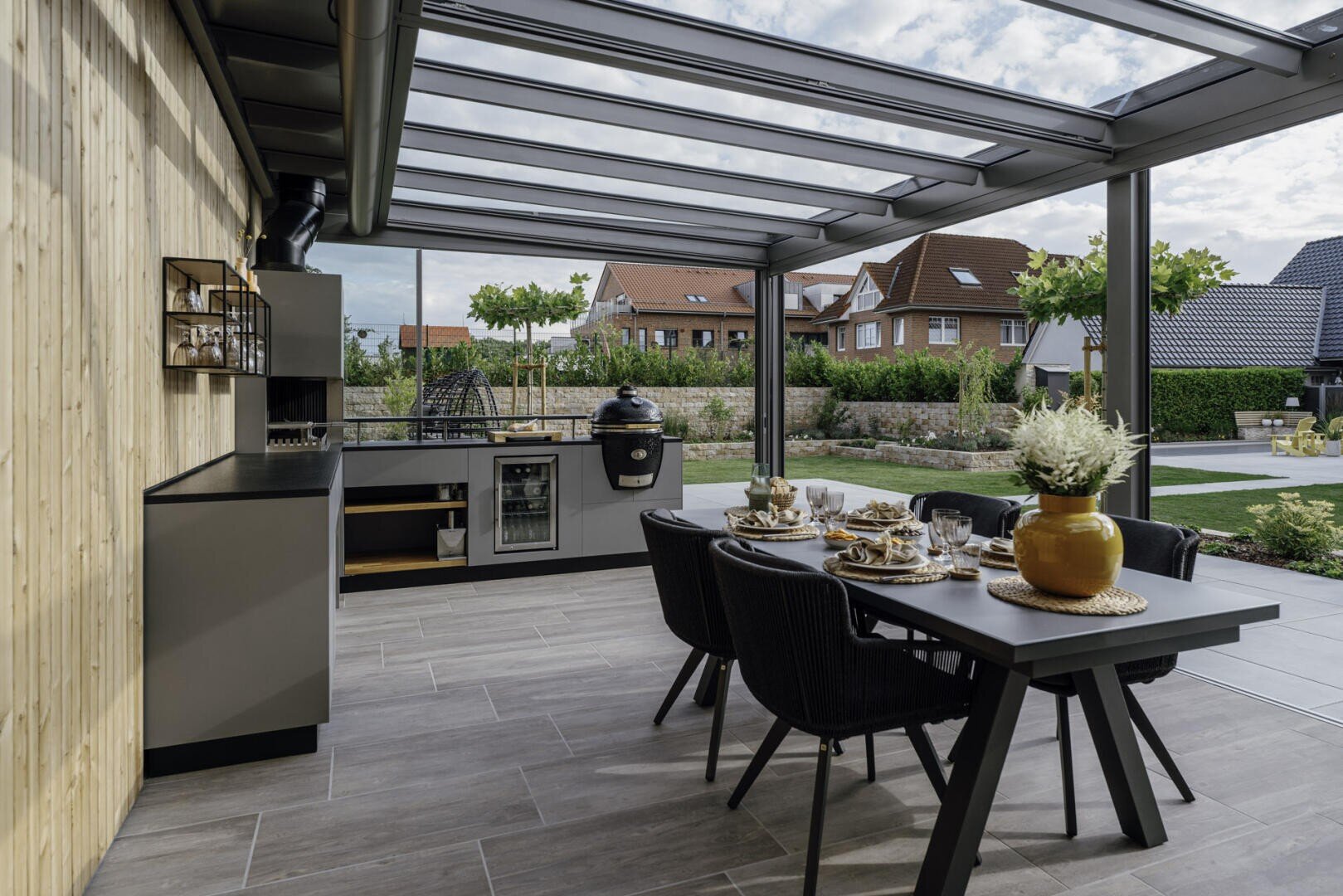 Moderne Außenküche und Essbereich unter einem Glasdach mit einem Esstisch für vier Personen, eingebautem Grill und Blick auf einen begrünten Hinterhof mit Häusern im Hintergrund.