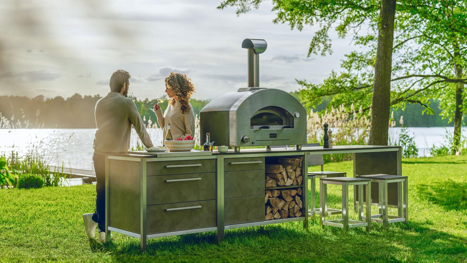 Ein Mann und eine Frau genießen gemeinsam Getränke an einem Pizzaofen und einer Kücheneinrichtung im Freien, umgeben von grünem Gras und Bäumen, mit einem See im Hintergrund unter einem teilweise bewölkten Himmel.