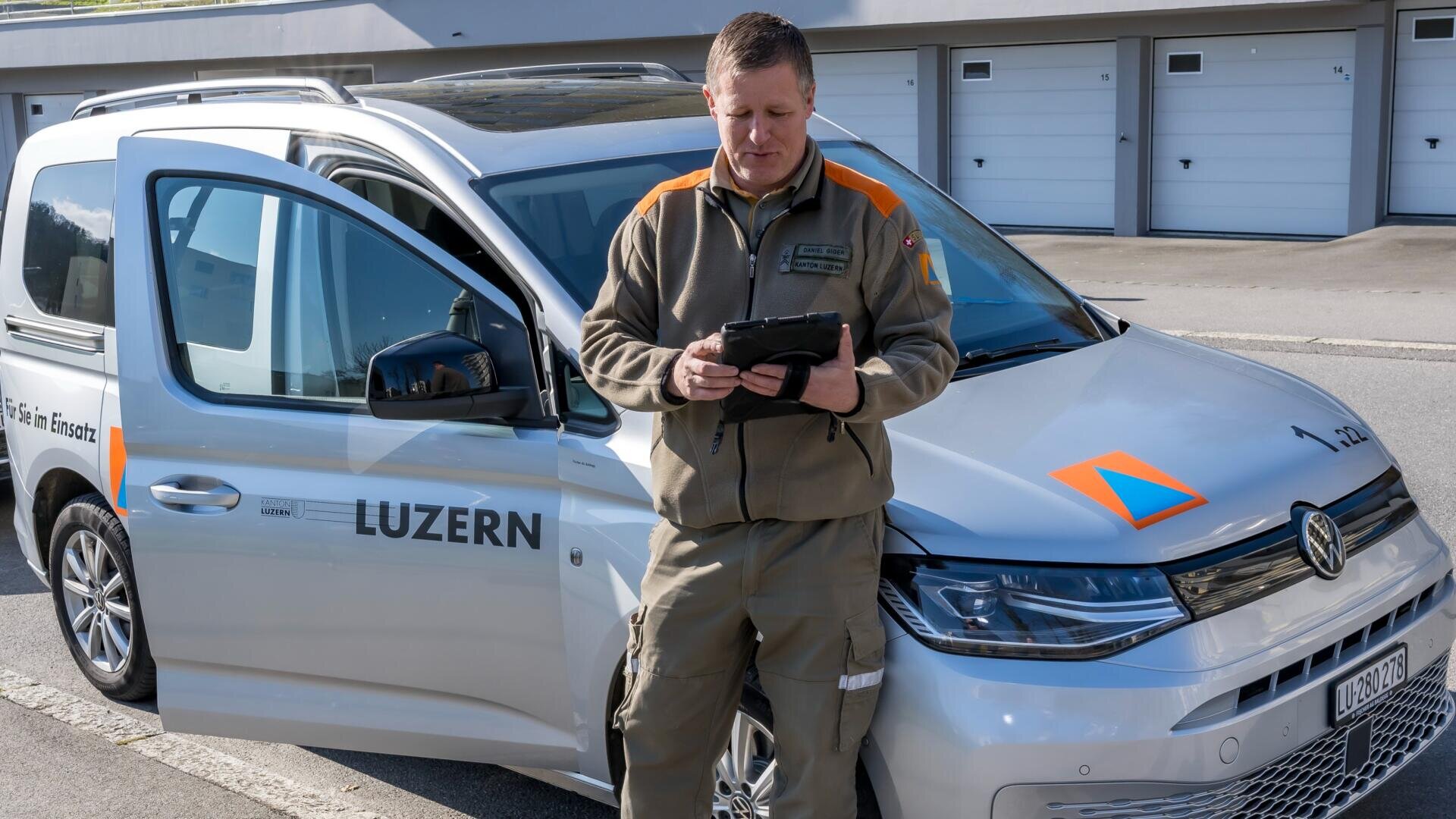 Ein Mann in Uniform steht vor einem silbernen Fahrzeug mit der Aufschrift LUZERN und benutzt ein Tablet. Die Autotür ist offen, und im Hintergrund sind Garagen zu sehen. Der Mann scheint zu arbeiten oder eine Inspektion durchzuführen.