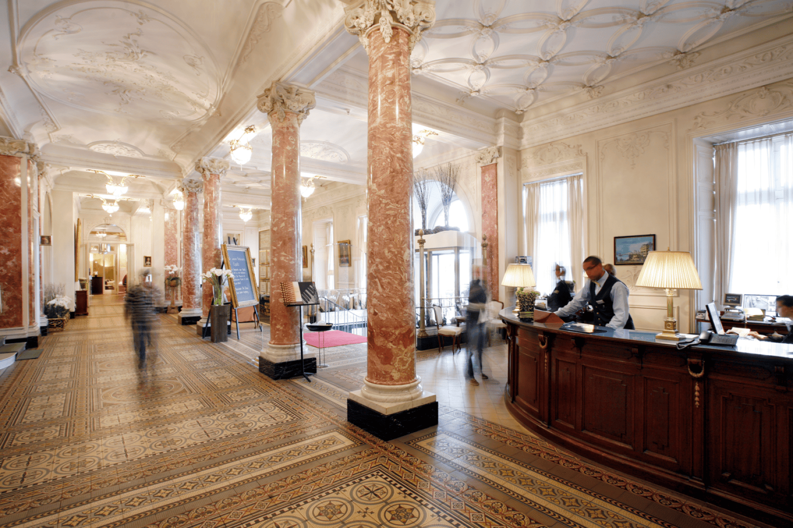 Große Hotellobby mit verzierten Decken, roten Marmorsäulen, gemustertem Fliesenboden und einem Empfangstresen aus dunklem Holz, an dem das Personal den Gästen behilflich ist; die Leute laufen durch den eleganten, gut beleuchteten Raum.