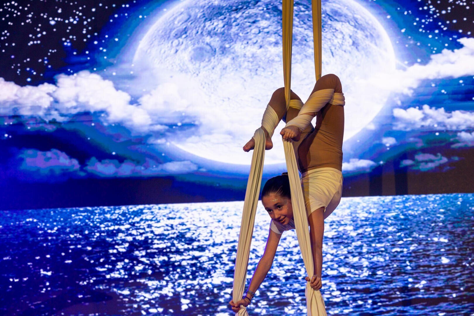 Eine Luftakrobatin in weißem Kostüm hängt kopfüber auf weißem Seidenstoff, mit einem großen beleuchteten Mond und einer Meeresszene im Hintergrund, die ein dramatisches und künstlerisches Bühnenbild schafft.