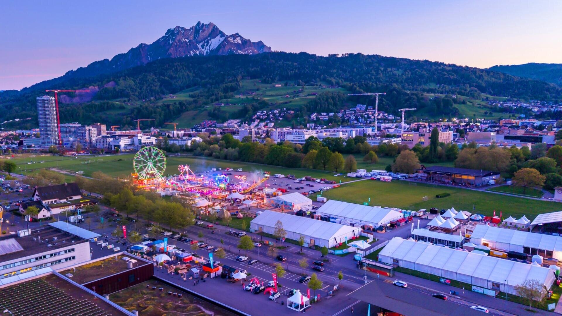 Luftaufnahme eines lebhaften Jahrmarkts mit Riesenrad, Zelten und bunten Lichtern vor dem Hintergrund einer Stadt, grüner Hügel und eines schneebedeckten Berges bei Sonnenuntergang.