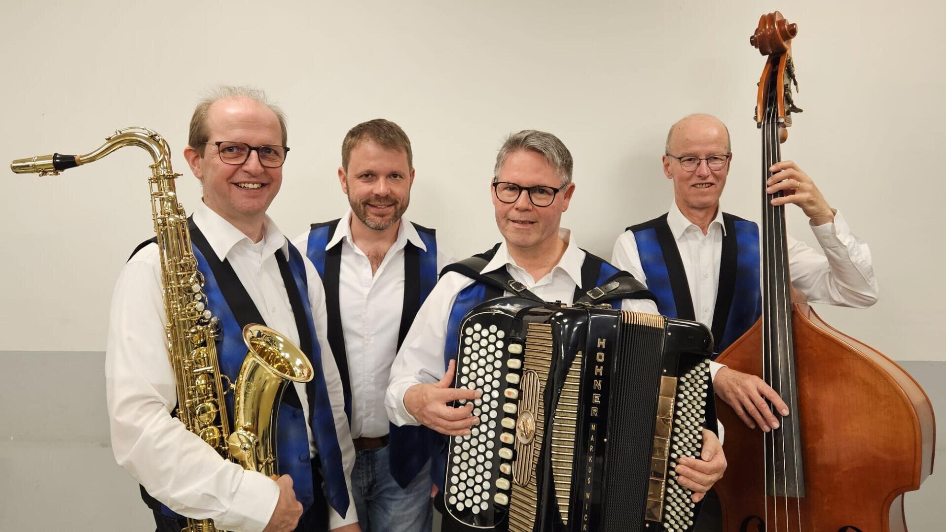 Vier Männer in weißen Hemden und blauen Westen stehen lächelnd mit Musikinstrumenten - einem Saxophon, einem Akkordeon und einem Kontrabass - vor einem schlichten hellen Hintergrund.