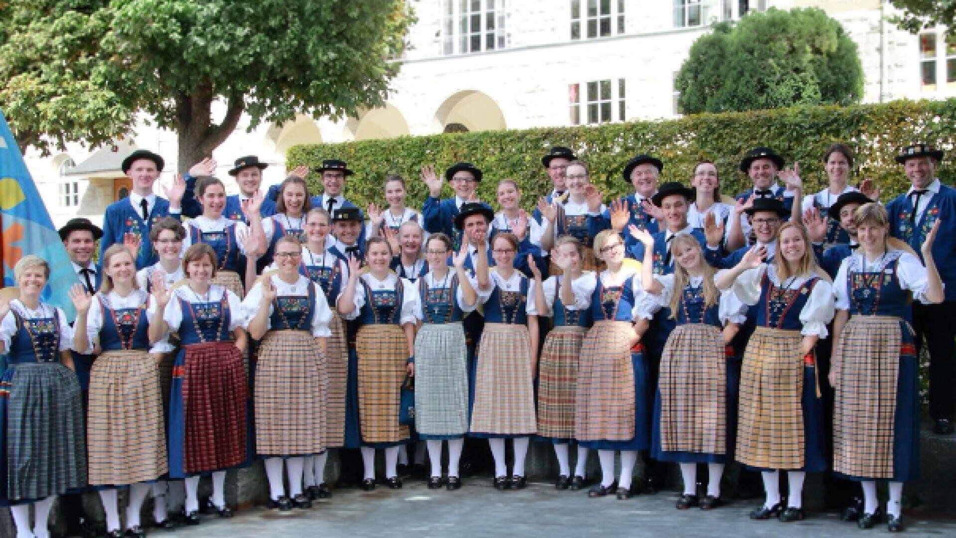 Eine große Gruppe von Menschen in traditioneller bayerischer Tracht steht im Freien, lächelt und winkt in die Kamera. Sie stehen in Reihen vor grünen Hecken und einem Gebäude mit Rundbogenfenstern.