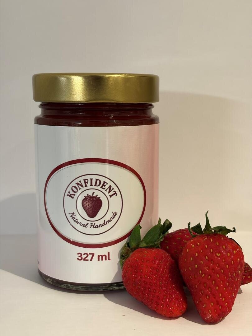 Ein Glas Konfident Natural Handmade Erdbeerkonfitüre mit goldenem Deckel, beschriftet mit 327 ml, steht neben drei frischen Erdbeeren auf weißem Hintergrund.