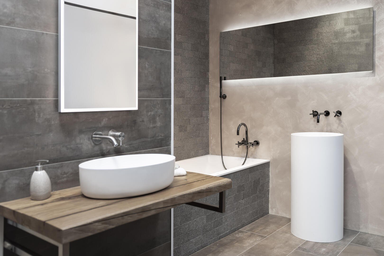 Modernes Badezimmer mit grau gefliesten Wänden, einem hölzernen Waschtisch mit rundem weißem Waschbecken, einem freistehenden zylindrischen Waschbecken, einer rechteckigen Badewanne und einem großen beleuchteten Spiegel über der Wanne.