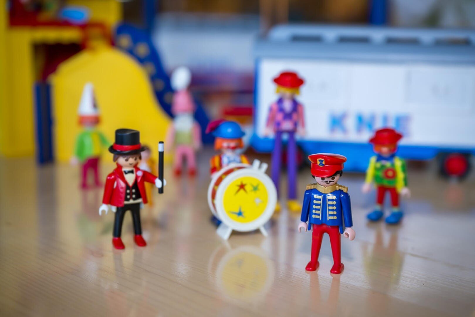 Bunte Spielzeugzirkusfiguren, darunter ein Zirkusdirektor, ein Clown und ein Trommler, stehen auf einer glatten Oberfläche mit einer Spielzeugtrommel und anderen verschwommenen Zirkusspielzeugen im Hintergrund.