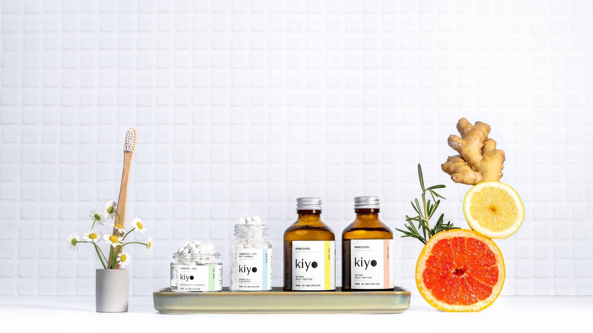 Hautpflegeprodukte mit der Aufschrift kiyo sind auf einem Tablett mit einer hölzernen Zahnbürste, Kamillenblüten, Ingwer, Zitrone, Grapefruit und Rosmarin vor einem weiß gekachelten Hintergrund angeordnet.