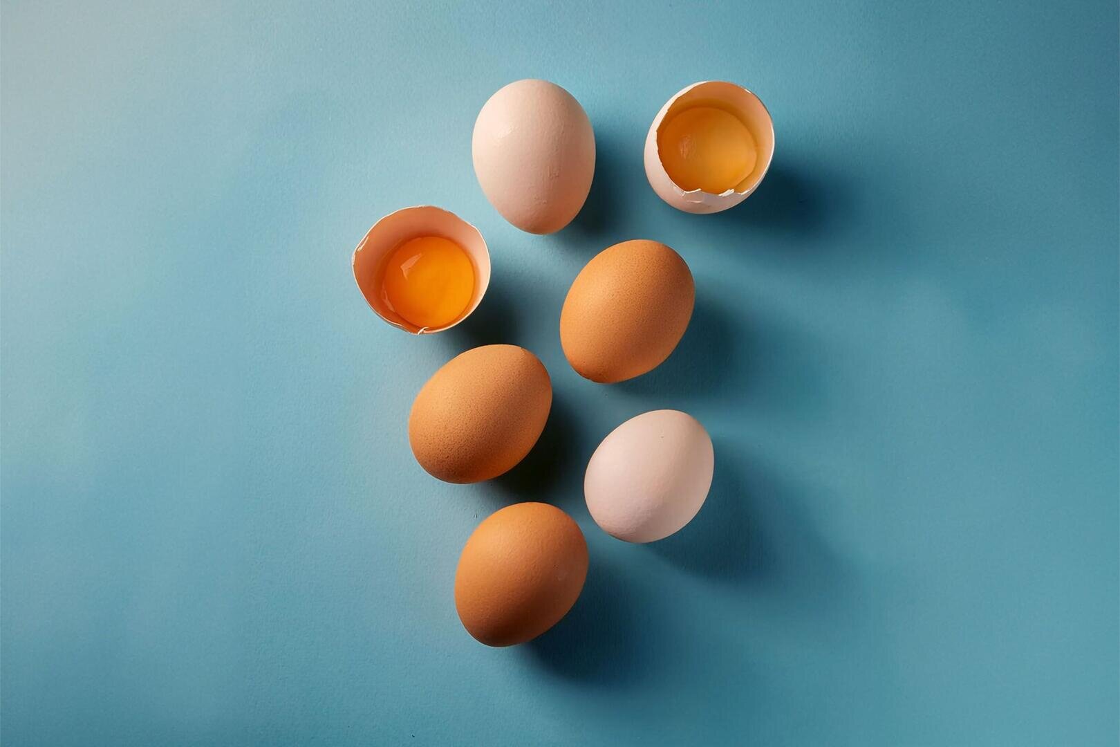 Sieben Eier, die auf einer blauen Fläche angeordnet sind, wobei zwei Eier aufgeschlagen sind und das Eigelb zeigen, und fünf ganze Eier (braun und weiß).