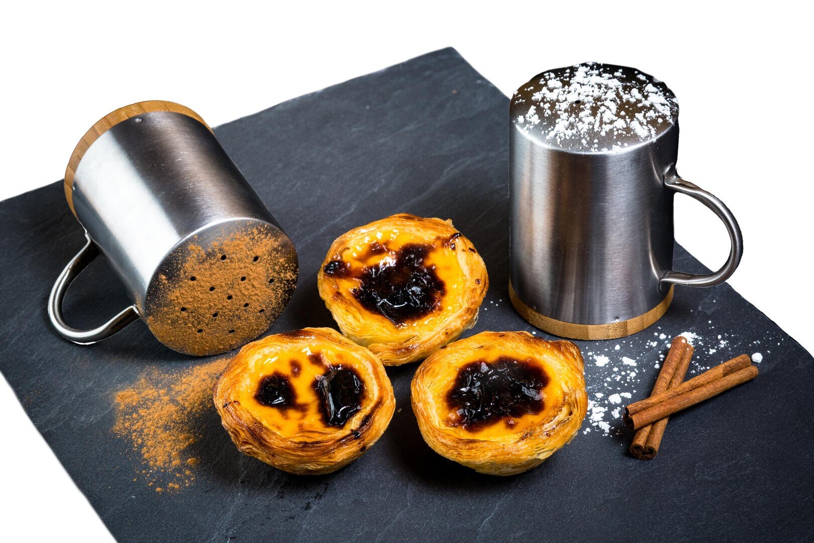 Drei portugiesische Puddingtörtchen auf einer Schiefertafel mit zwei Metallstreuern - einer mit Zimt, einer mit Puderzucker - und zwei Zimtstangen, alles auf weißem Hintergrund.