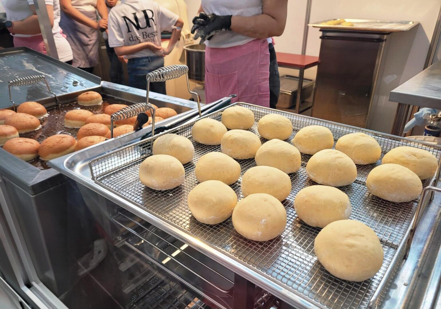 Teigkugeln liegen auf einem Metallgestell neben einer Fritteuse mit goldbraunen Brötchen, während im Hintergrund Menschen auf einem Markt oder an einem Essensstand stehen.