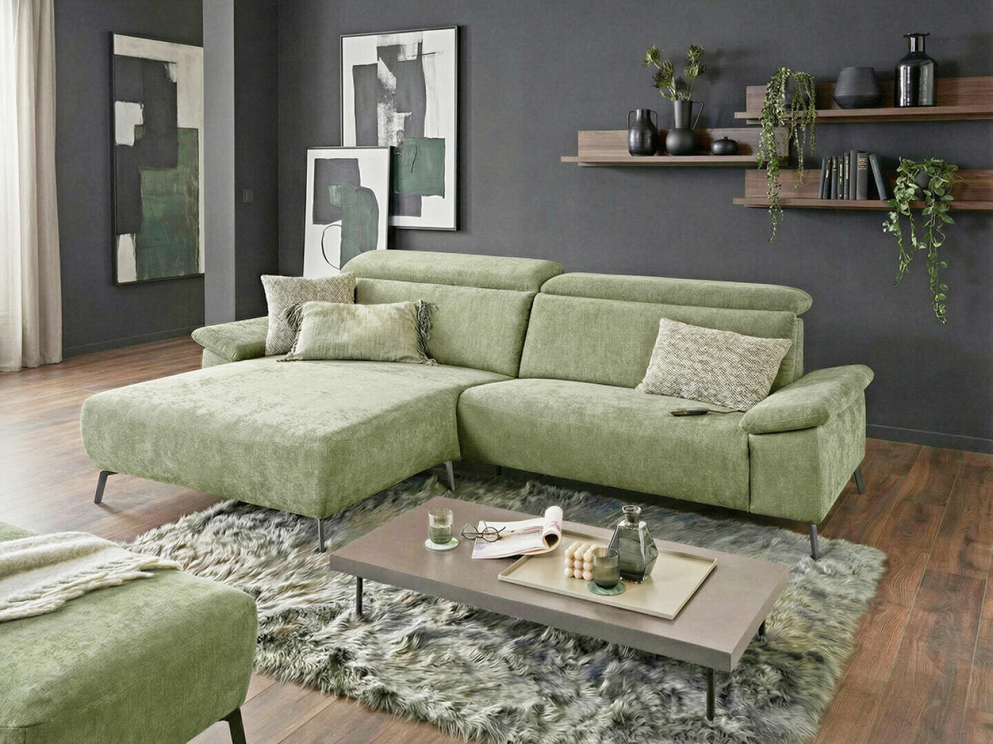 Ein modernes Wohnzimmer mit einem hellgrünen Sektionssofa, strukturierten Kissen, einem weichen grauen Zottelteppich und einem niedrigen rechteckigen Couchtisch. Dunkelgrüne abstrakte Kunst und Pflanzen schmücken die dunklen Wände und schwebenden Regale.
