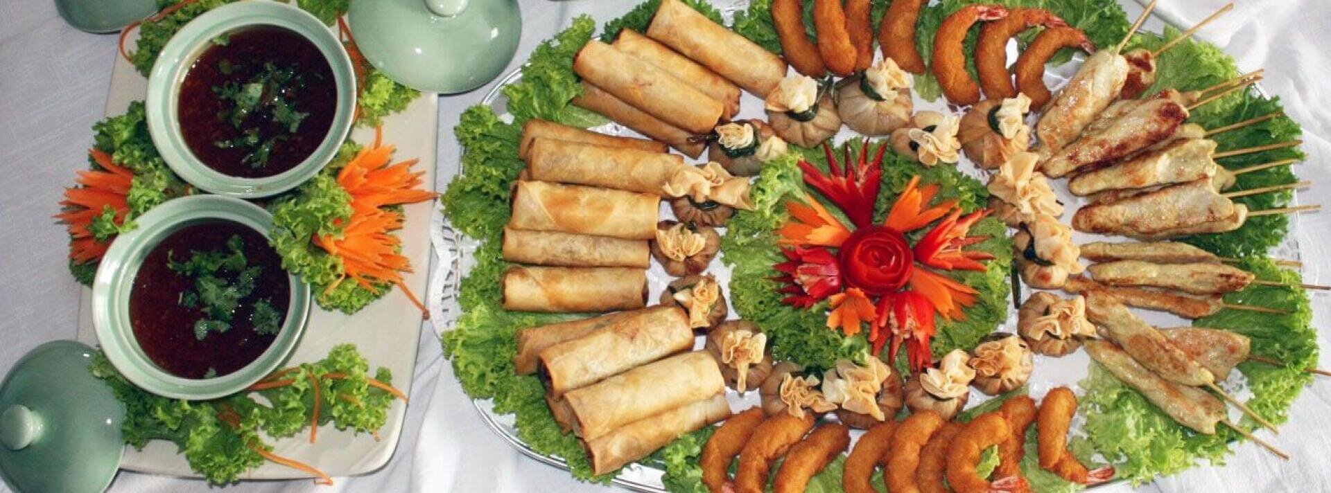 Eine Platte mit verschiedenen asiatischen Vorspeisen, darunter Frühlingsrollen, gebratene Teigtaschen, Spieße und Garnelen, ist auf einem Salatteller mit einem geschnitzten Gemüsestück angerichtet. Zwei Schalen mit Dip-Sauce mit Kräutern stehen neben der Platte.