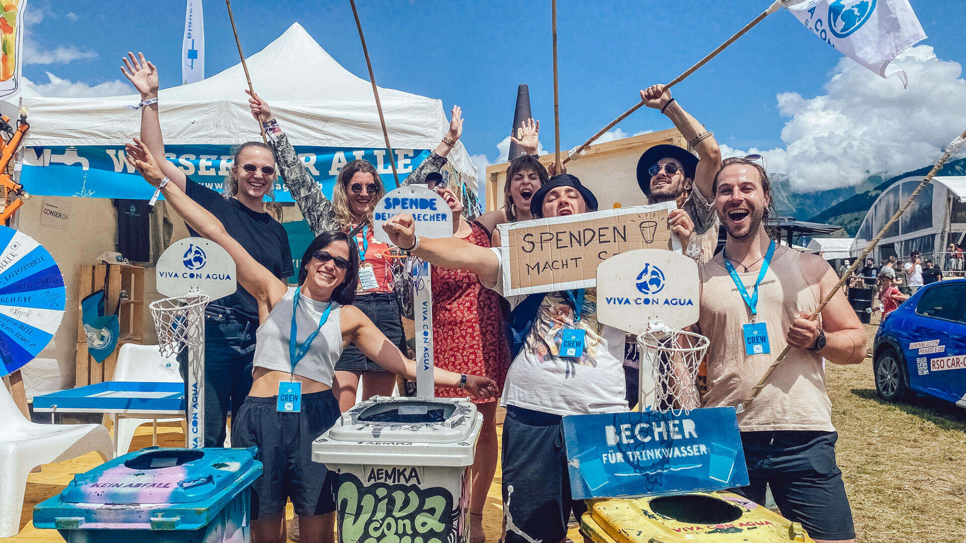 Eine Gruppe lächelnder Menschen posiert im Freien an einem Festivalstand mit Recycling-Tonnen und Spendenschildern und hebt feierlich die Arme. Im Hintergrund sind der blaue Himmel und die Berge zu sehen.