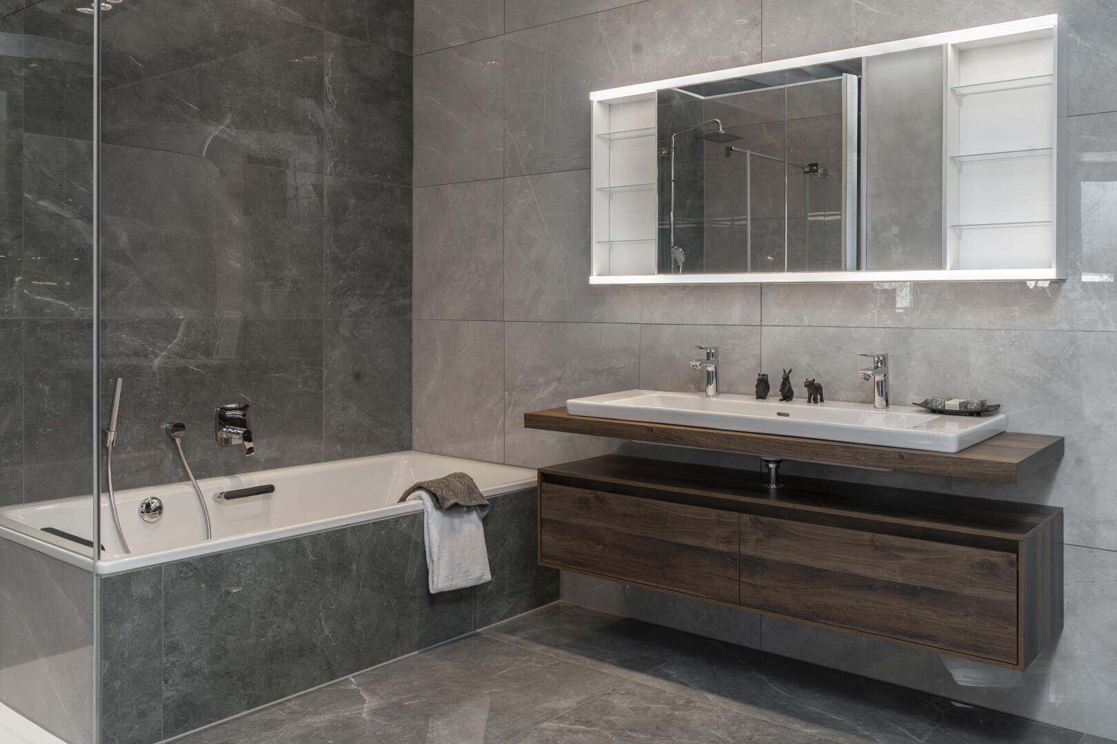 Modernes Badezimmer mit grauen Marmorfliesen, einer eingebauten Badewanne mit Handtuch, einem schwebenden hölzernen Waschtisch mit Doppelwaschbecken, zwei Wasserhähnen und einem großen beleuchteten Spiegel mit Ablageflächen auf beiden Seiten.