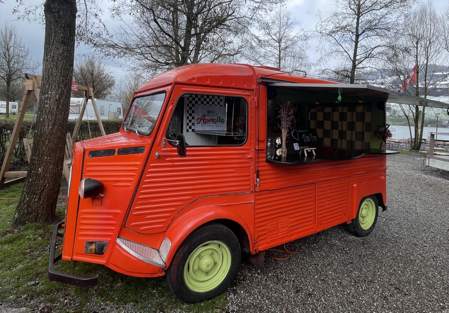 Ein leuchtend roter Foodtruck mit grünen Rädern ist auf einer Kiesfläche in der Nähe von Bäumen und einem Fluss geparkt. Das Servicefenster ist geöffnet und auf einem Schild im Inneren steht "Acapella".