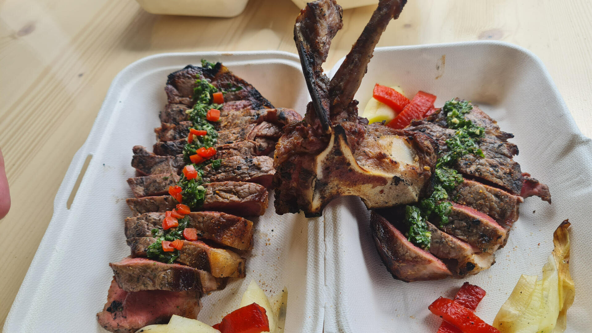 Zwei Portionen gegrilltes Fleisch mit Knochen, übergossen mit grüner Chimichurri-Sauce und gewürfelter roter Paprika, serviert in einem Behälter zum Mitnehmen mit geröstetem Gemüse auf einem Holztisch.