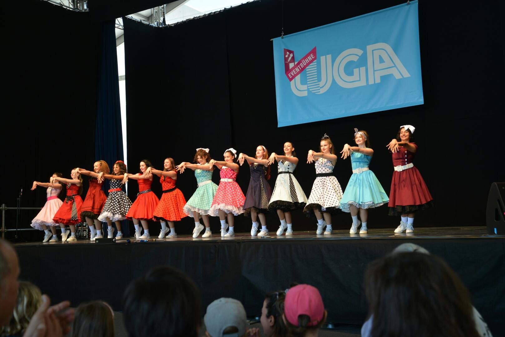 Eine Gruppe von Mädchen in farbenfrohen Kleidern im Vintage-Stil führt auf der Bühne vor einem Publikum einen choreografierten Tanz auf, während im Hintergrund ein großes blaues Banner mit der Aufschrift UGA zu sehen ist.