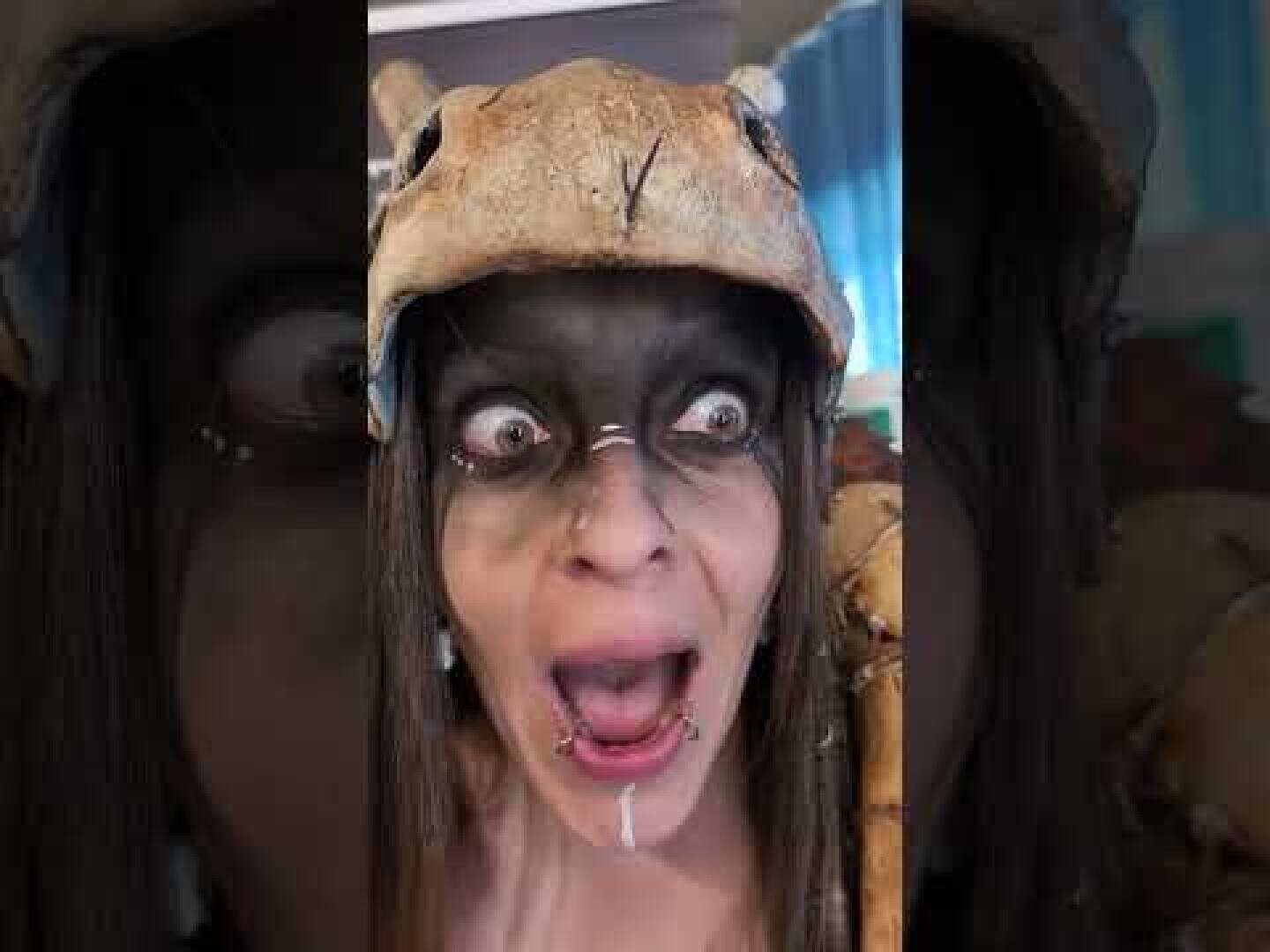 Eine Person mit großen Augen und offenem Mund trägt Gesichtsbemalung und einen Hut, der einem Tierschädel ähnelt. Ihr Gesicht ist mit schwarzen und weißen Zeichen bemalt, und sie hat Piercings an den Lippen und der Nase.