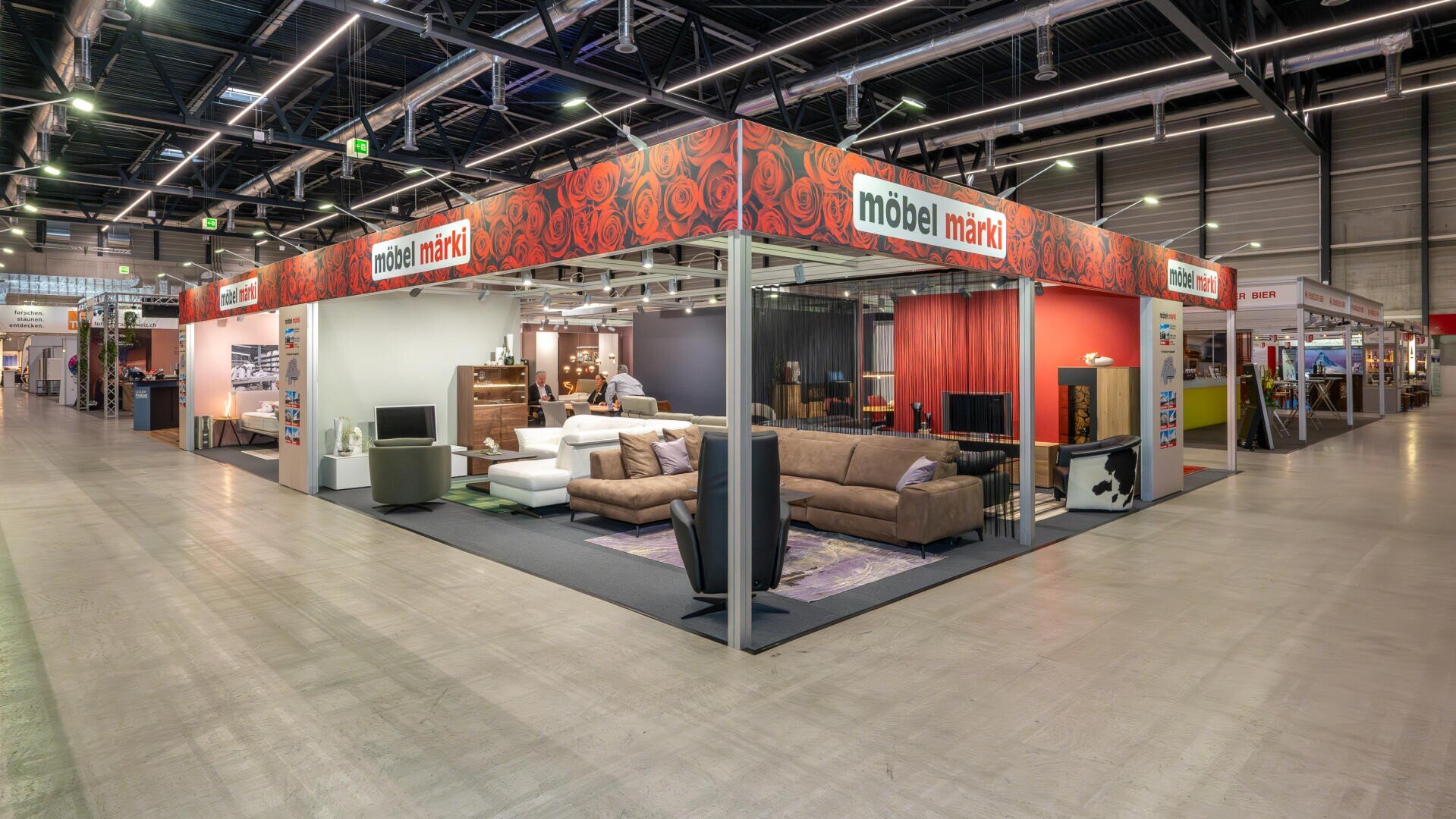 Ein Stand eines Möbelhauses auf einer Indoor-Messe zeigt Sofas, Stühle, Tische und Regale in hellem Licht. Der Stand hat eine rote Beschilderung mit dem Namen "Möbel Märki" und dekorativen Blumenmustern auf den oberen Platten.