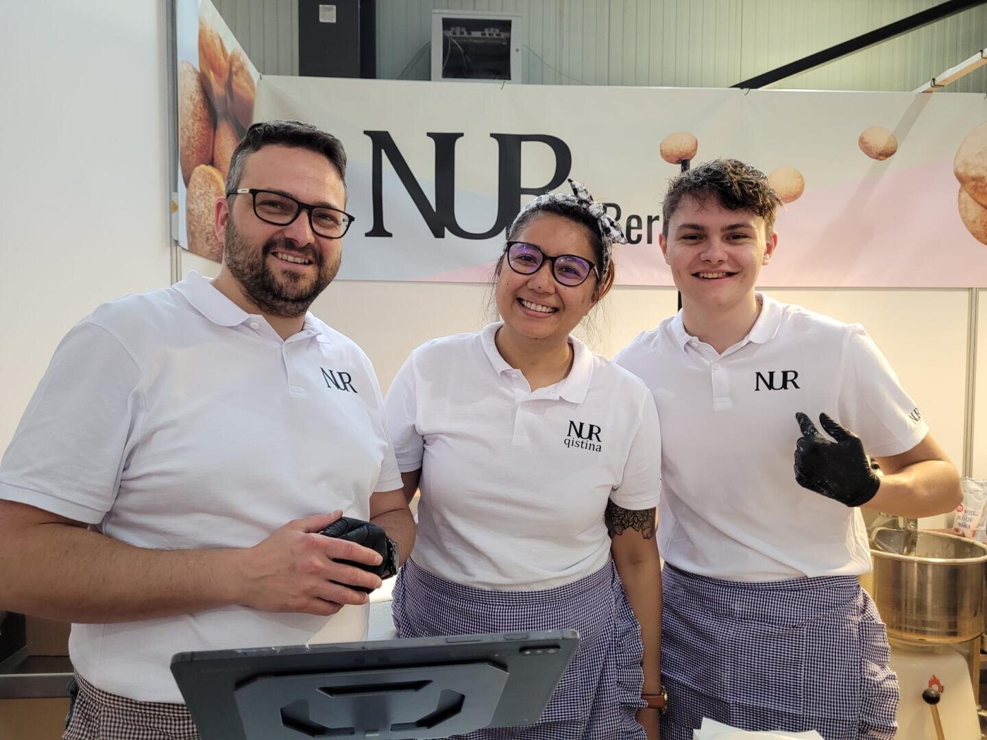 Drei Personen in weißen NR-Hemden und karierten Schürzen lächeln hinter einer Theke in die Kamera. Möglicherweise handelt es sich um einen Essensstand oder einen Marktstand mit einem NR-Schild im Hintergrund.