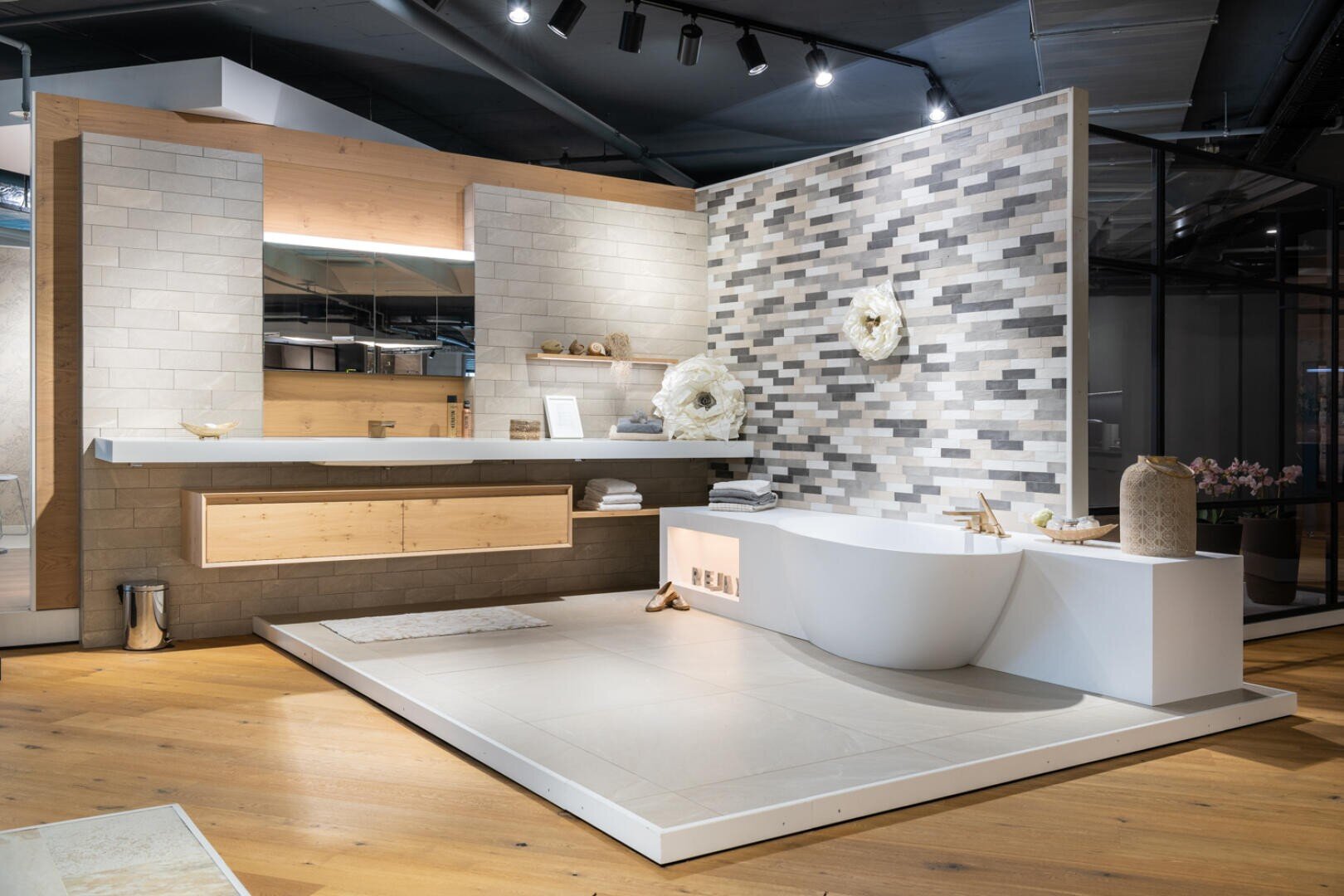 Ein modernes Badezimmer mit einer freistehenden weißen Badewanne, einem schwebenden Waschtisch und einer Wand mit grauen und beigen Fliesen. Der Raum ist mit Holzböden, minimalistischem Dekor und weichen, neutralen Tönen ausgestattet.