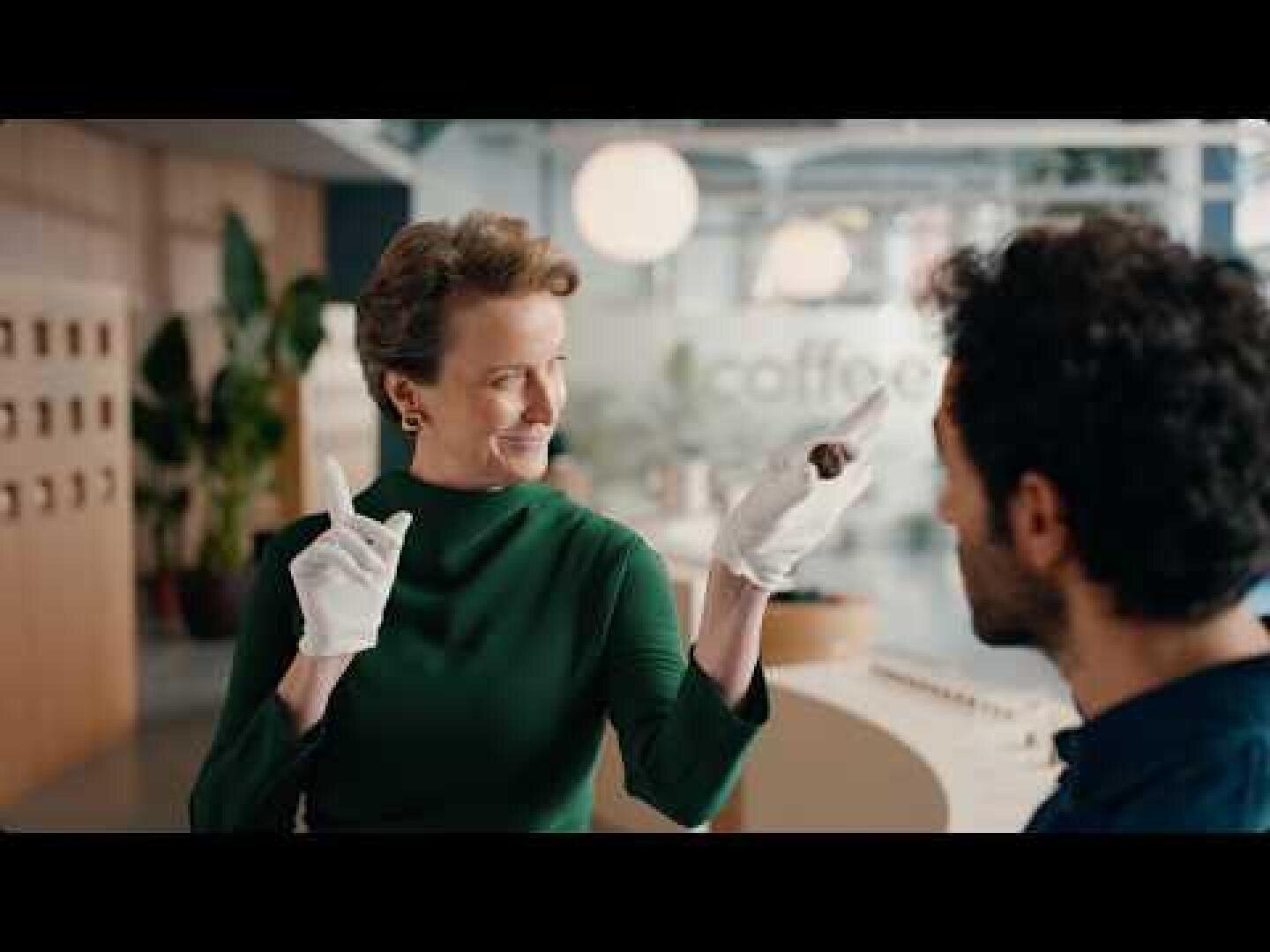 Eine Frau in einem grünen Kleid und weißen Handschuhen lächelt, während sie einen kleinen Gegenstand hochhält und nach oben zeigt. Sie steht in einem hellen, modernen Raum mit Pflanzen und blickt auf einen sitzenden Mann, der der Kamera den Rücken zuwendet.