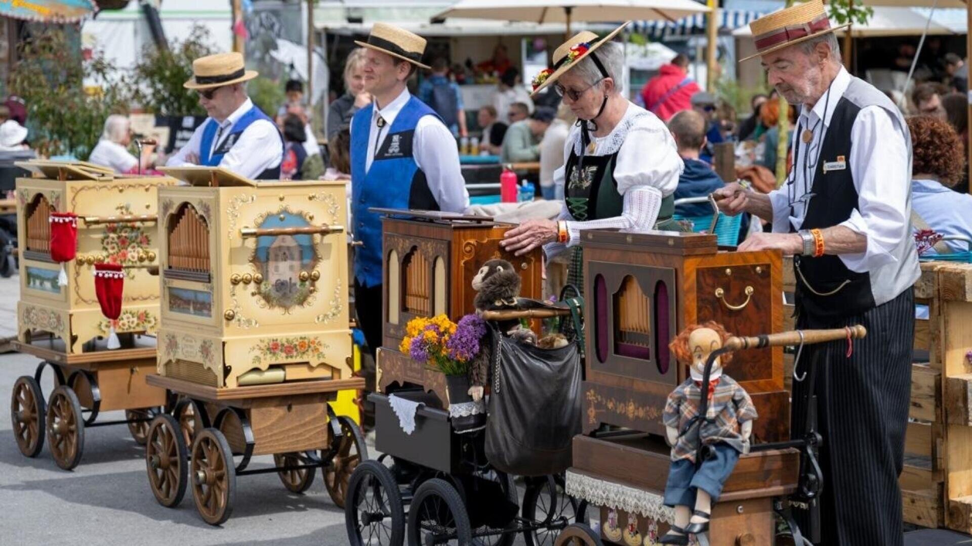 Vier traditionell gekleidete Personen spielen bei einer Veranstaltung im Freien auf einem Rollwagen eine verzierte Drehorgel. Die Instrumente sind mit Puppen, Blumen und komplizierten Mustern verziert. Im Hintergrund sitzen Menschen.