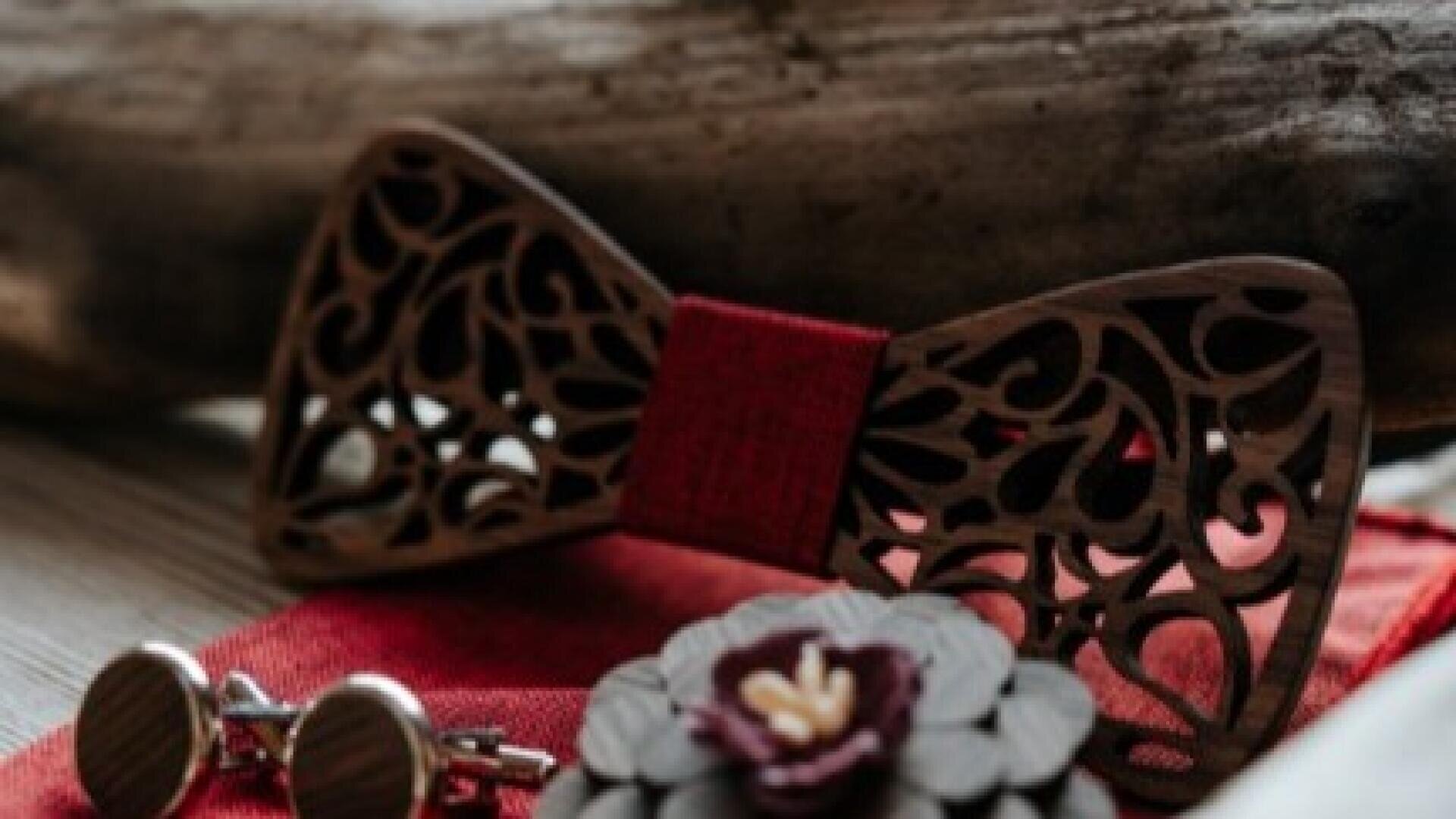 Eine hölzerne Fliege mit komplizierten Ausschnittmustern und einer roten Mitte, gepaart mit hölzernen Manschettenknöpfen und einer blumenförmigen Reversnadel, arrangiert auf einem roten Tuch.