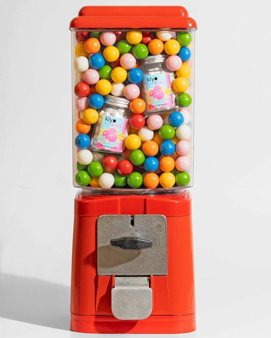 Ein roter Kaugummiautomat, gefüllt mit bunten runden Kaugummis, darunter rosa, gelb, grün, blau, orange und weiß, mit zwei kleinen Gläsern mit Süßigkeiten zwischen den Kaugummis.