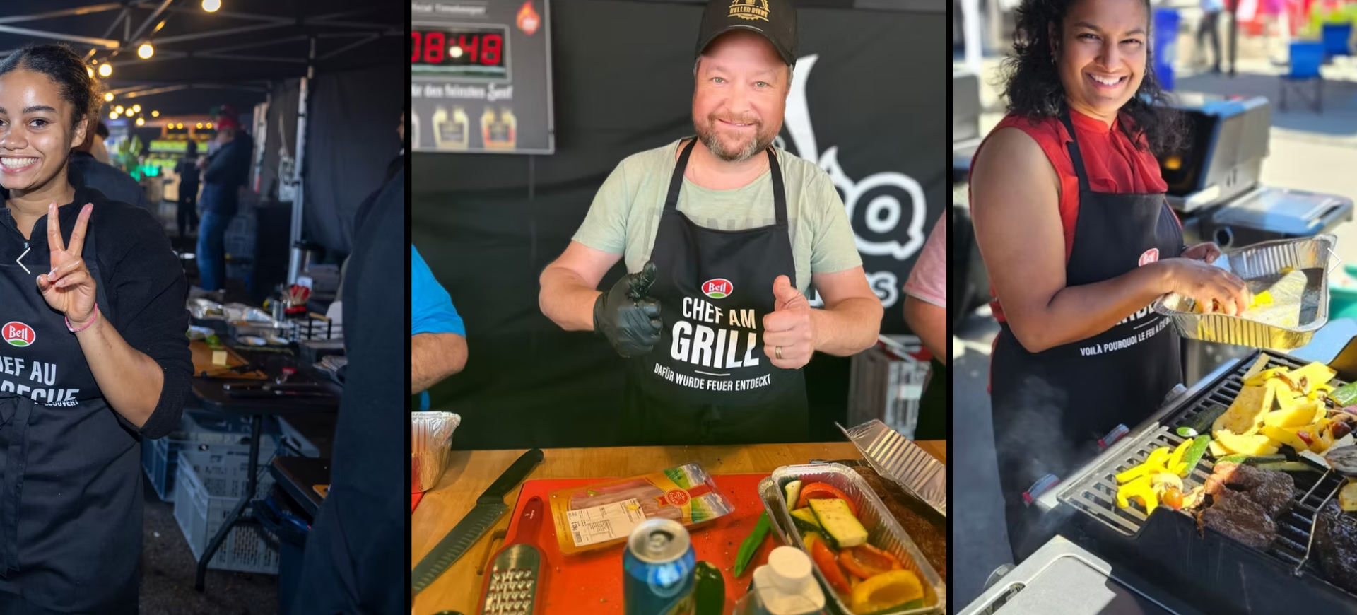 Drei Personen mit Schürzen lächeln, während sie bei einer Veranstaltung im Freien grillen. Eine Frau macht ein Friedenszeichen, ein Mann zeigt den Daumen nach oben, und eine andere Frau kocht Gemüse und Fleisch auf einem Grill.