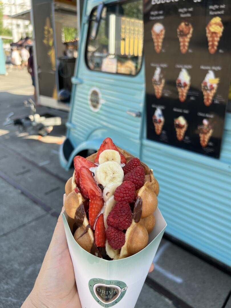 Eine Hand hält eine Waffeltüte mit Erdbeerscheiben, Bananen, Himbeeren und Schokoladensoße vor einem blauen Imbisswagen mit einer Speisekarte, auf der verschiedene Desserts stehen.