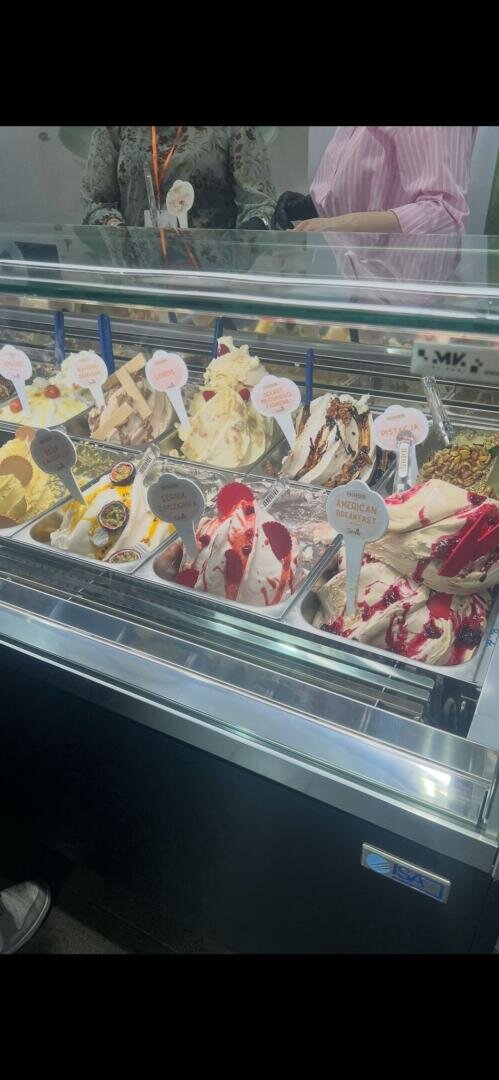 Eine Vitrine mit verschiedenen Gelatine-Sorten, die mit bunten Strudeln und Garnierungen versehen sind. Geschmacksschilder sind zu sehen, und zwei Personen stehen hinter der Theke, teilweise sichtbar über dem Glas.