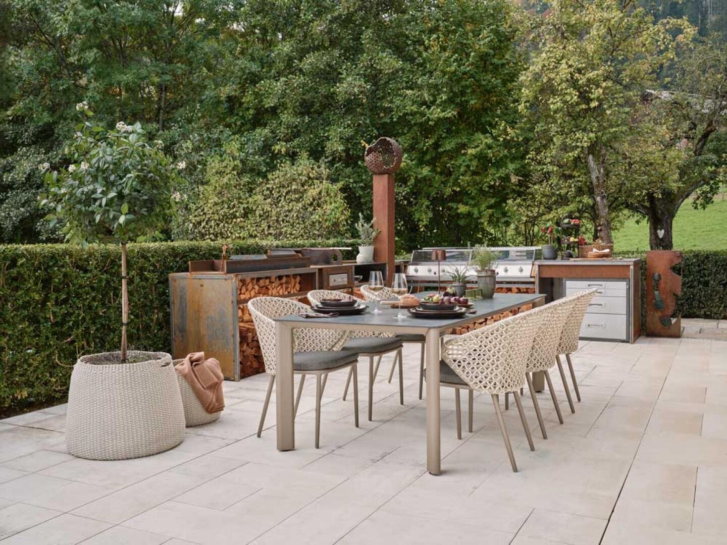 Eine Außenterrasse mit einem modernen Esstisch für sechs Personen, umgeben von geflochtenen Stühlen. Im Hintergrund befinden sich ein eingebauter Grill und ein Küchenbereich mit aufgestapeltem Brennholz, umgeben von grünen Bäumen und Hecken.
