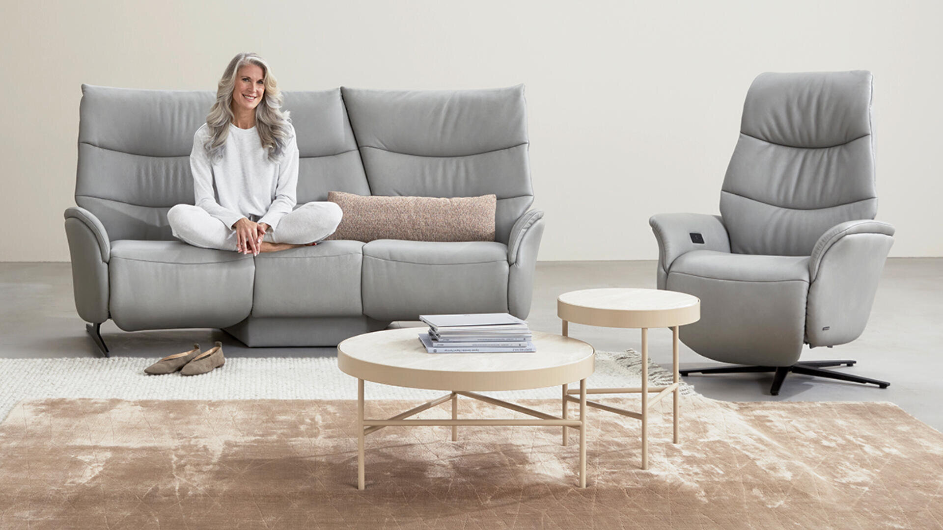 Eine Frau mit langen grauen Haaren und in heller Kleidung sitzt im Schneidersitz auf einem modernen grauen Sofa. Ein passender Ruhesessel, runde Couchtische und ein neutralfarbener Teppich vervollständigen die minimalistische Wohnzimmereinrichtung.