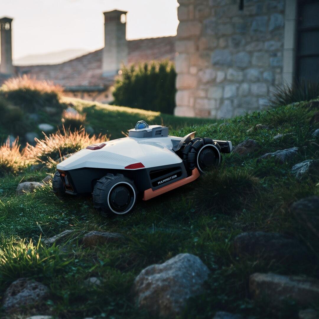 Ein Rasenmähroboter mit großen Rädern fährt im frühen Morgen- oder späten Nachmittagssonnenlicht einen grasbewachsenen, felsigen Hang in der Nähe eines Steinhauses hinauf.