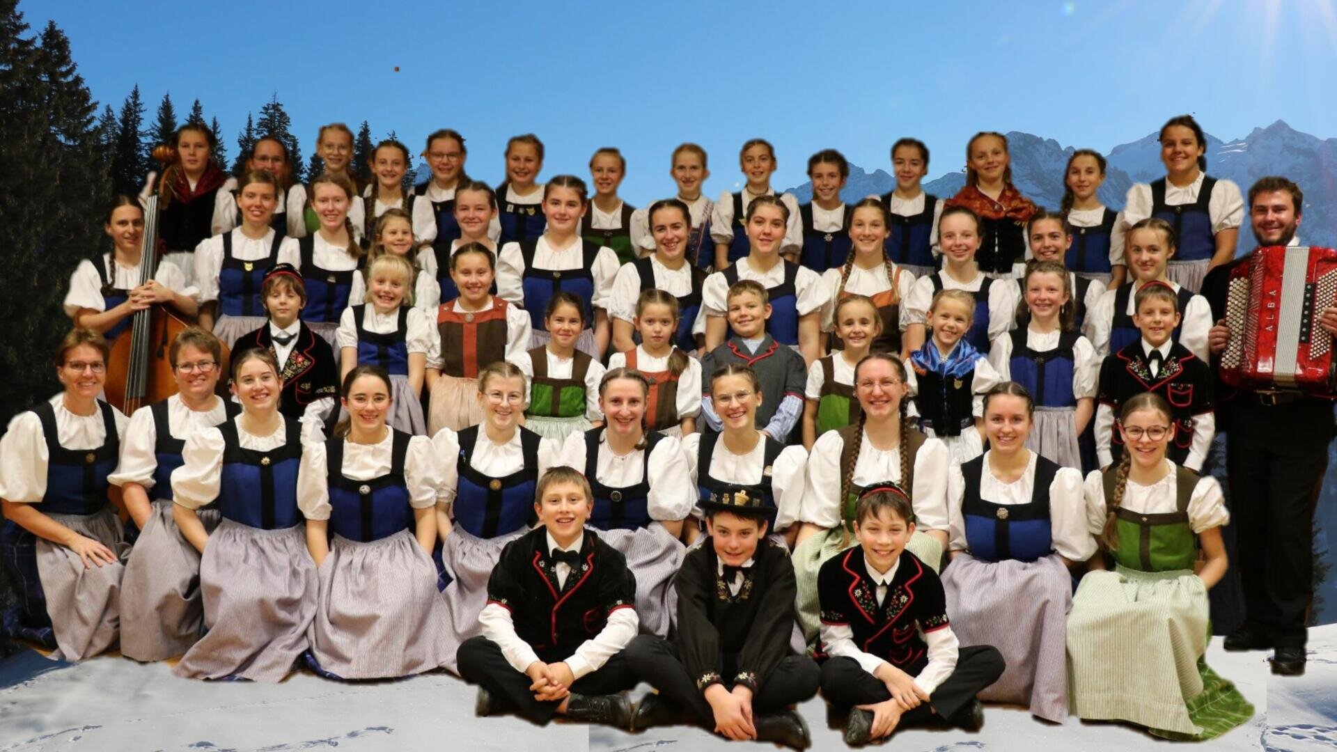 Eine große Gruppe von Kindern und Erwachsenen in traditioneller bayerischer Tracht posiert für ein Gruppenfoto in einem Innenraum, vor dem Hintergrund von Bergen, Bäumen und blauem Himmel. Auf der rechten Seite steht ein Akkordeonspieler.
