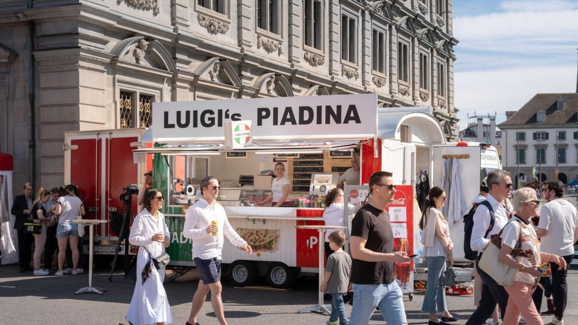 Ein Imbisswagen namens Luigis Piadina bedient Kunden bei einer Veranstaltung im Freien. Mehrere Menschen gehen vorbei oder stehen herum, einige mit Essen und Getränken in der Hand, mit einem historischen Gebäude im Hintergrund.