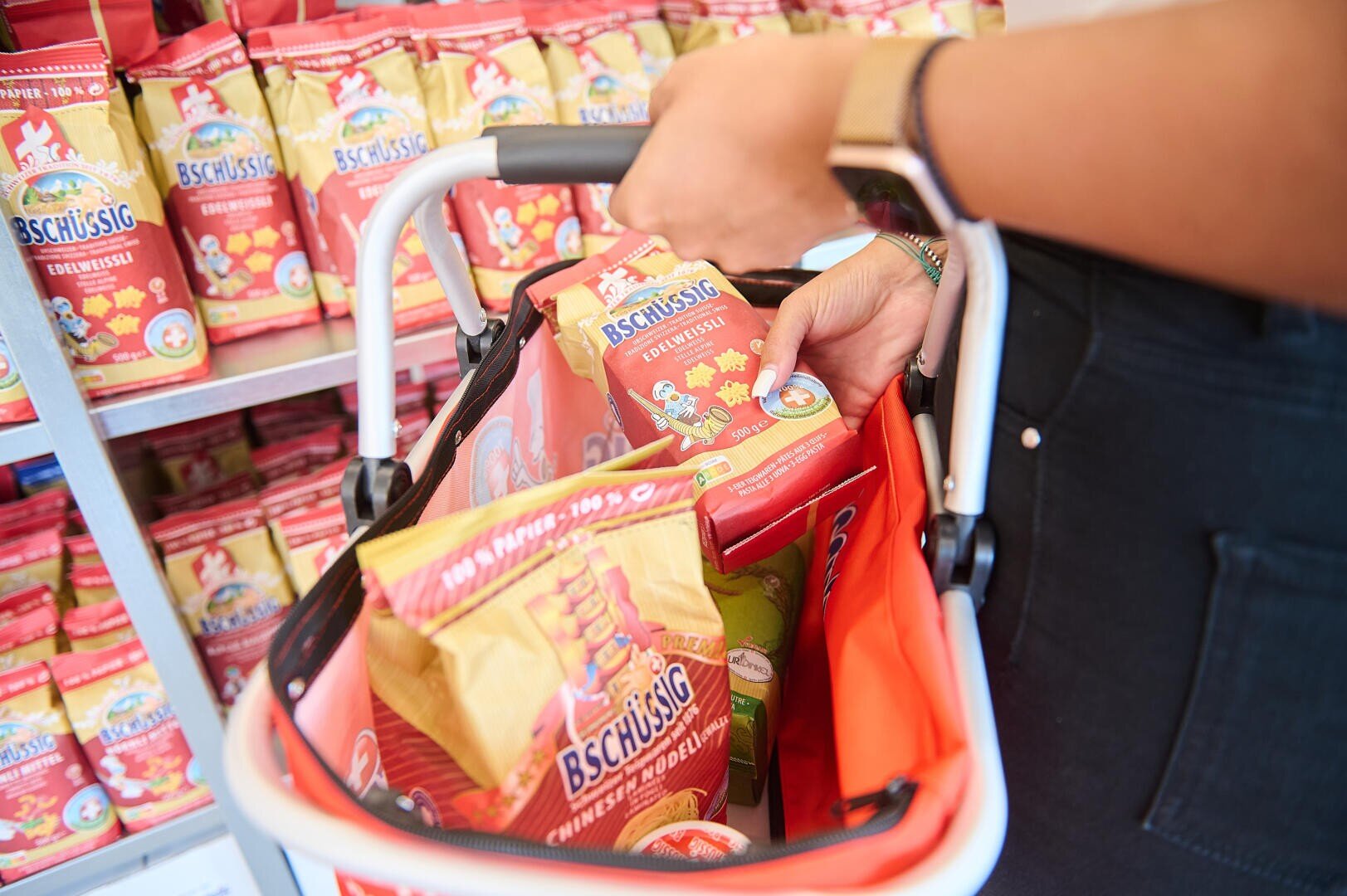Eine Person legt eine Packung Nudeln der Marke Bschüssig in einen Einkaufskorb, in dem sich bereits andere Bschüssig-Produkte befinden, vor ein Regal mit ähnlichen Artikeln.