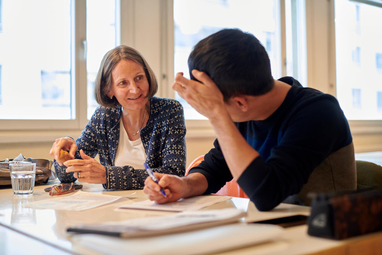 Eine Frau lächelt, während sie sich mit einem Mann unterhält, der neben ihr an einem Tisch sitzt. Sie scheinen zusammen zu arbeiten, mit Papieren, Stiften und einem Glas Wasser vor sich in einem hell erleuchteten Raum.