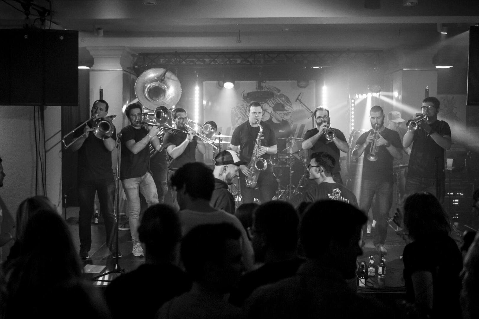 Schwarz-Weiß-Foto einer Blaskapelle auf der Bühne mit neun Musikern, die Posaunen, Trompeten und ein Sousaphon spielen. Vorne steht ein Publikum, das die energiegeladene Live-Performance verfolgt.