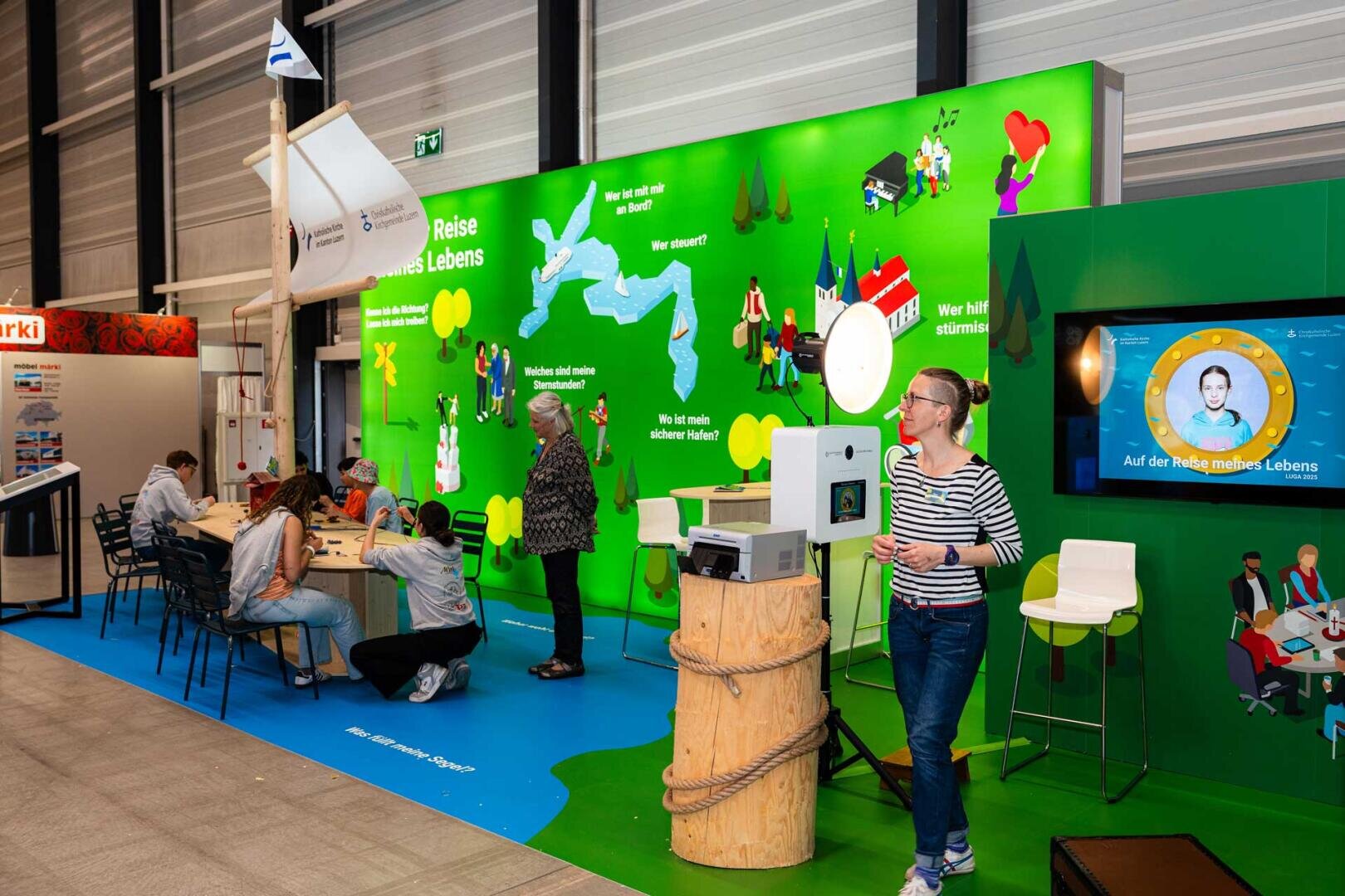 In einem hell erleuchteten Raum mit grünen Wänden, auf denen bunte Illustrationen und Texte mit Fragen zu sehen sind, befinden sich Menschen. Eine Frau steht neben einer Fotobox mit Bildschirm, während andere an einem Tisch sitzen oder stehen und sich mit Aktivitäten beschäftigen.