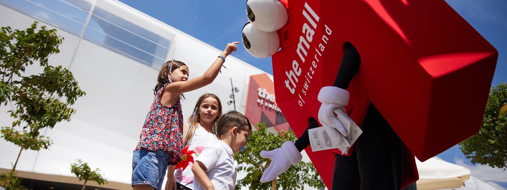 Drei Kinder interagieren an einem sonnigen Tag im Freien in der Nähe eines modernen Gebäudes mit einem großen roten Maskottchen, das wie eine Einkaufstasche gekleidet ist, auf der das Einkaufszentrum der Schweiz steht.