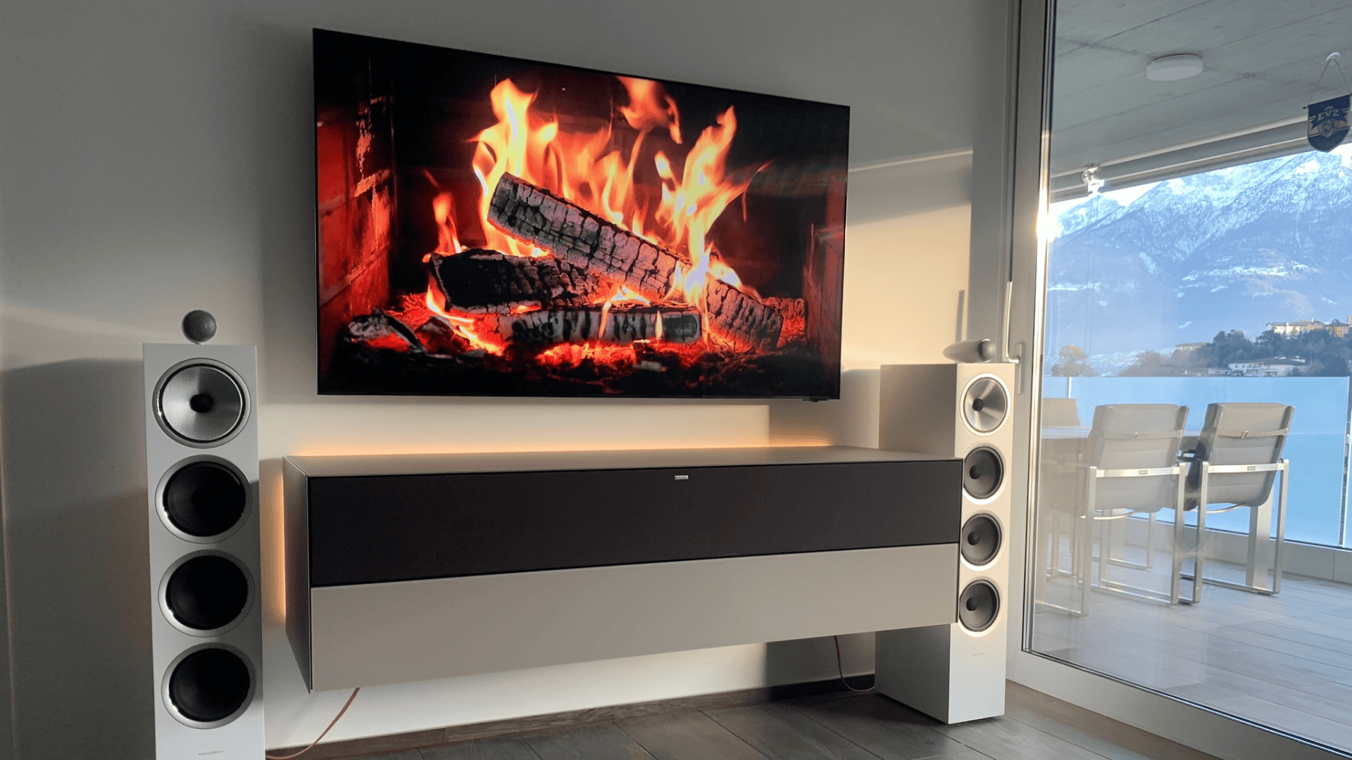 Ein modernes Wohnzimmer mit einem an der Wand montierten Fernseher, der einen brennenden Kamin zeigt, flankiert von zwei weißen Bodenlautsprechern. Große Fenster geben den Blick frei auf die Berge und einen See mit einem Tisch und Stühlen im Freien.