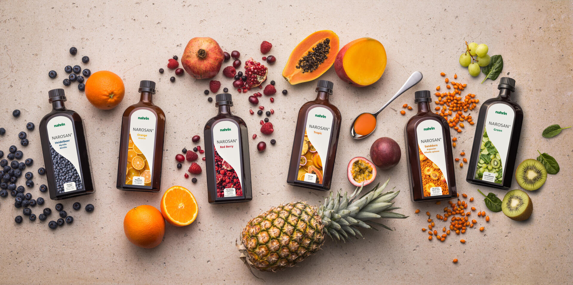 Sechs Flaschen Fruchtsirup sind auf einer Fläche angeordnet, jede umgeben von den wichtigsten Früchten: Blaubeeren, Orangen, Granatäpfel, Papaya, Ananas, Passionsfrucht, Sanddorn und Kiwi, zusammen mit geschnittenem Obst und Sirup.