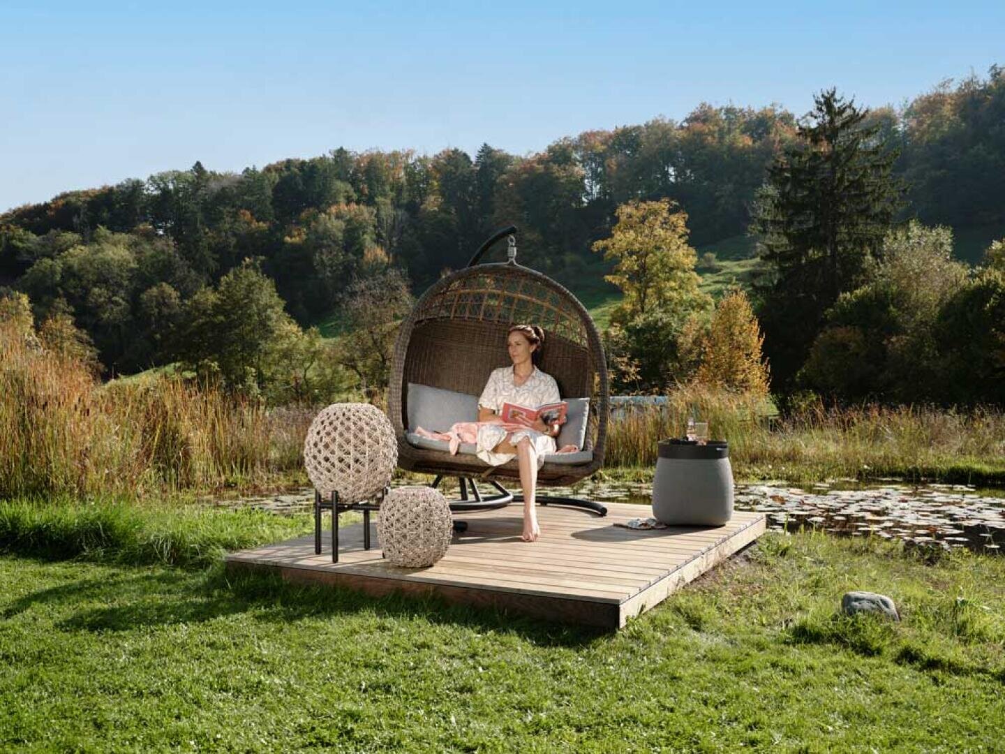 Eine Frau sitzt in einem großen, runden Gartenstuhl auf einer Holzplattform an einem Teich und liest eine Zeitschrift, umgeben von Grün und Bäumen unter einem klaren blauen Himmel. In der Nähe stehen kugelförmige Dekorationsgegenstände aus Weidengeflecht.